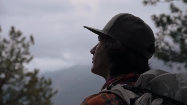 Крупным планом видно молодого длинноволосого парня в кепке, выходящего вечером из леса в облачную погоду в горах. Треккинг и путешествия — стоковое видео