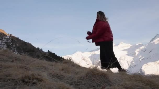 Attraktiv ung kvinna i svart klänning och röd dunjacka går uppför en gräsbevuxen sluttning mot de snöklädda höga bergen i Kaukasus — Stockvideo