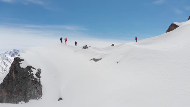 Bir grup kayakçı ve snowboardcunun yüksek tepelerin arka planına karşı karla kaplı bir dağın tepesindeki hava manzarası. Kayakçılardan biri dağ yamacının dik yamacında sürmeye başlar. — Stok video