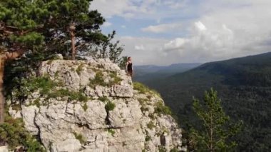 Siyah tişörtlü ve şortlu genç bir adam yaz mevsiminde bir uçurumun kenarında bir orman dağı vadisine karşı durur. Hava görünümü