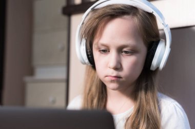 Yüzü kulaklıklı üzgün küçük bir kız yerde dizüstü bilgisayarla oturuyor ve ödevini yapıyor. Uzak öğrenme
