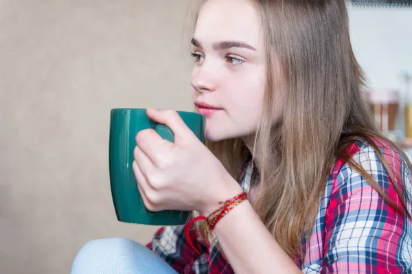 Jovem mulher sorrindo atraente bebe chá ou café de uma caneca grande na cozinha e olha para o lado — Fotografia de Stock