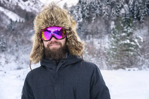 Retrato de um atraente homem barbudo sorriso em uma máscara de esqui e um chapéu de pele contra o fundo de uma floresta nevada — Fotografia de Stock