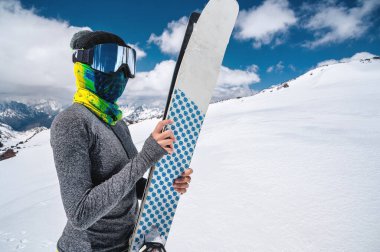 Dağlarda kar maskesi ve türban takmış ince bir kayakçı duruyor. Kayakları karın arka planında tutar