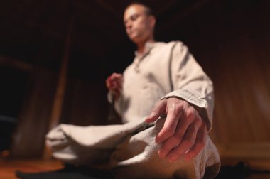 Gri keten elbiseli bir adam karanlık bir meditasyon odasında lotus pozisyonunda oturur ve elinde kırmızı bir tespihle meditasyon yapar..