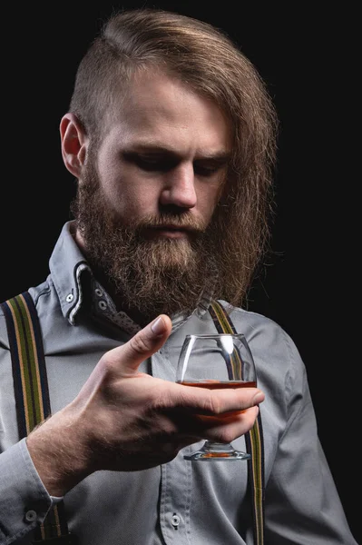 Elegante hombre barbudo en una camisa con tirantes huele un vaso de whisky. retrato de estudio — Foto de Stock