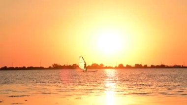 Acemi bir rüzgar sörfçüsü kadın günbatımı arka planında yelkenliyle sörf tahtasının üzerinde duruyor. Rüzgar sörfü okulu
