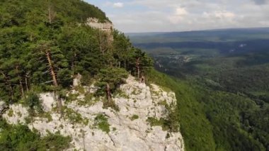 Uzun saçlı, siyah tişörtlü ve şortlu zayıf bir adam uçurumun kenarında oturur ve etrafındaki orman ve dağlara bakar. Hava görünümü
