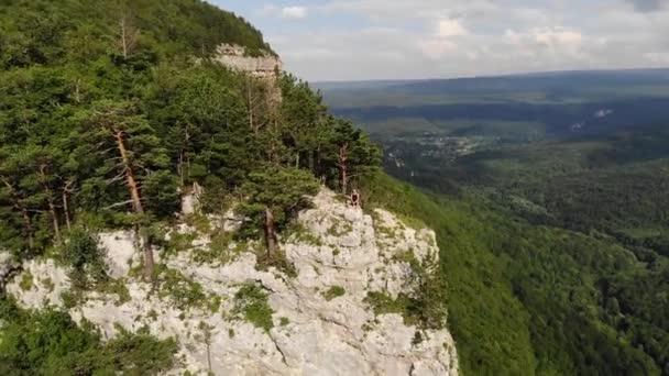 Ein langhaariger, dünner Mann in schwarzem T-Shirt und kurzer Hose sitzt am Rande einer Klippe und blickt auf den umliegenden Wald und die Berge. Luftaufnahme — Stockvideo