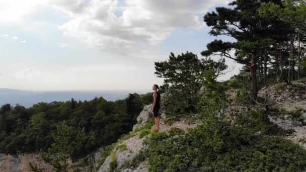 Ein langhaariger, hagerer Mann in schwarzem T-Shirt und kurzer Hose steht am Rande einer Klippe und blickt auf den umliegenden Wald und die Berge. Luftaufnahme — Stockvideo