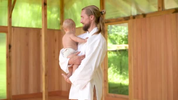 Um pai, um praticante de ioga e qigong em roupas soltas de cor clara, brinca com sua filha de um ano de idade no salão de prática. Segura-a e gira-a — Vídeo de Stock