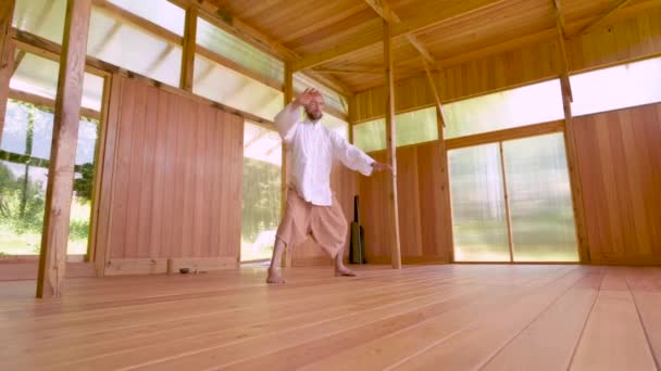 En långhårig vit man i lätt och lös klädsel övar qigong tai chi i ett övningsrum i trä på sommaren. Långsamma rörelser harmoni och lugn — Stockvideo