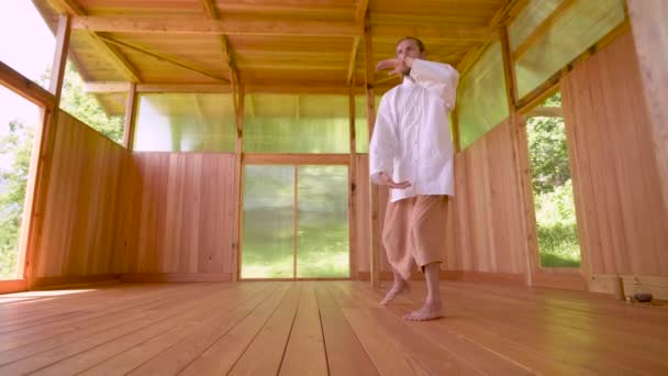 Een langharige blanke man in lichte en losse kleding oefent 's zomers qigong tai chi in een houten oefenruimte. Langzame bewegingen harmonie en rust — Stockvideo