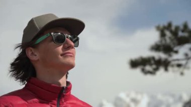 Uzun saçlı, kırmızı ceketli ve güneş gözlüklü gri şapkalı bir gencin portresi, uzaktaki bir dağ geçidinde arka planda karla kaplı zirvelerle kameraya bakıyor.