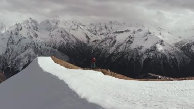 Sırt çantalı güneş gözlüklü genç adam kar ve taşlardan oluşan tepenin tepesinden kar örtülü dağlara doğru iniyor..