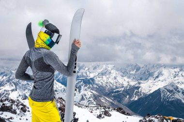 Tüylü ince bir kızın portresi kar maskeli kar maskeli bir kar maskeli bir kar maskesi ve kar örtülü destansı dağların arka planında kayakların yanındaki kapalı yüzlü bir şapka..