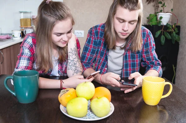 A young smile couple guy and a girl with long hair sit at the dinner table in the kitchen and use phones communicating in chat rooms and social networks. — Stock Photo, Image