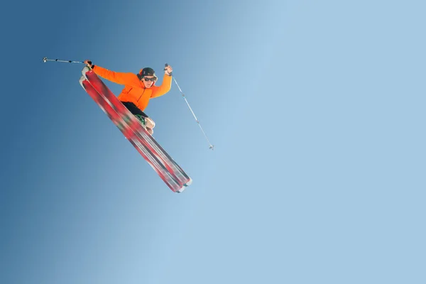Stylischer Skifahrer mit orangefarbener Jacke und Sonnenbrille im Sprung macht den Trick isoliert auf blauem Hintergrund — Stockfoto