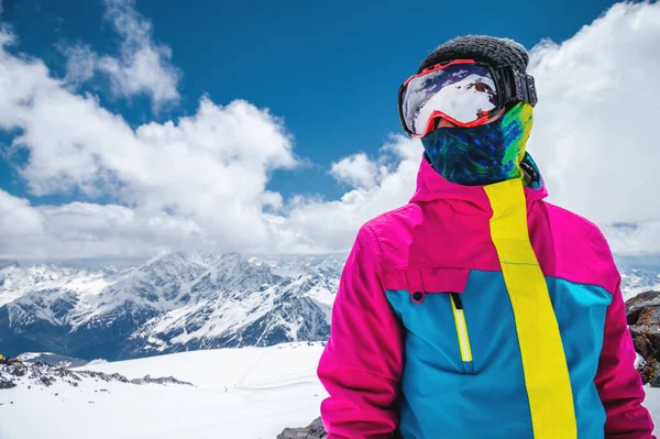Portrét dívčího lyžaře v pestrobarevné svěží bundě v lyžařské masce s zakrytou tváří za slunečného dne na pozadí zasněžených hor a mraků — Stock fotografie
