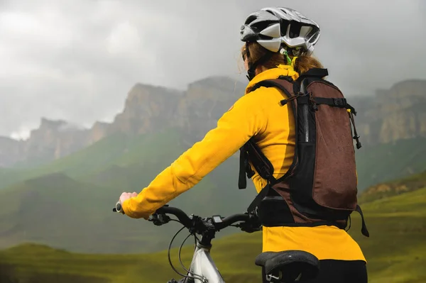 Primer plano de una joven esbelta con una mochila y un casco de bicicleta se sienta en una bicicleta alta en las montañas en un día nublado y mira a las montañas y las nubes. —  Fotos de Stock