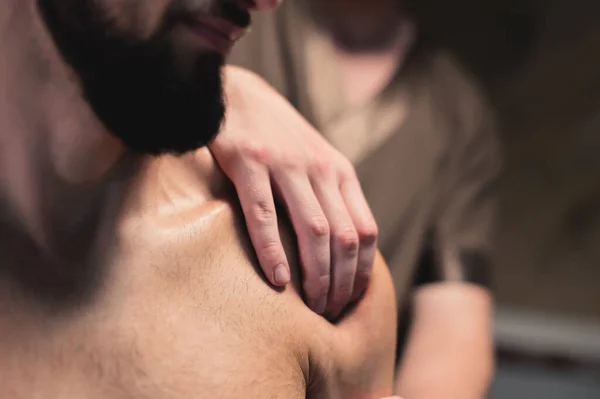 Masér zblízka provádí sportovní masáž ramen svalnatému sportovci v místnosti s kontrastním tmavým světlem. Profesionální sportovní masáž — Stock fotografie