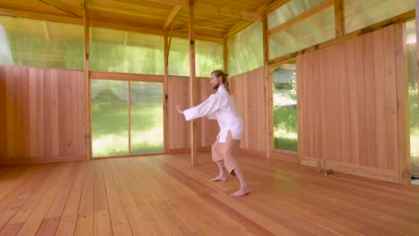 A long-haired Caucasian man in light and loose clothing practices qigong tai chi in a wooden practice room in summer. Slow movements harmony and calm — Stock Video