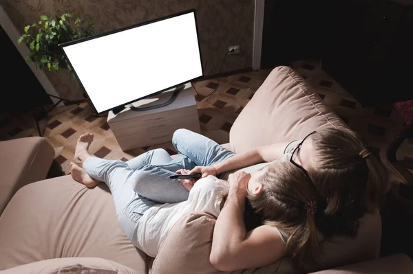Young family of long haired man and woman are lying on the couch and watching TV. The TV screen is cut out white. Mockup and template for designer layout