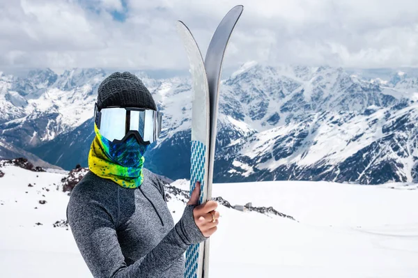 Um esquiador esbelto com uma máscara de esqui e um lenço de cabeça fica nas montanhas. segura esquis e olha para a câmera — Fotografia de Stock