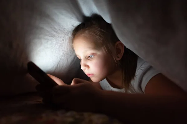 Маленька дівчинка вночі зі смартфоном під обкладинками. Дитяча залежність від гаджетів і безсоння — стокове фото