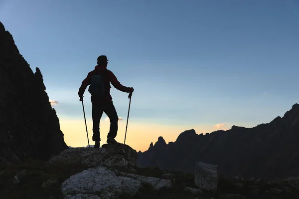 Silueta mladého muže v čepici a slunečních brýlích s batohem a trekkingovými tyčemi stojí večer vysoko v horách na pozadí hřebene. — Stock fotografie