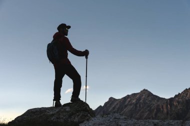 Sırt çantası ve yürüyüş direkleri olan şapkalı bir adamın silueti, akşam karanlığında karanlık gökyüzünün arka planına karşı, dağlarda yüksekte duruyor. Alçak tuş
