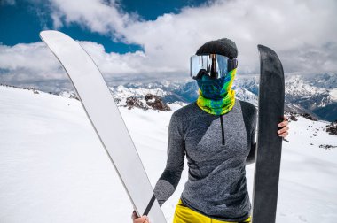 Dağlarda kar maskesi ve türban takmış ince bir kayakçı duruyor. Kayakları karın arka planında tutar