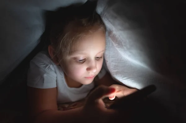 Geceleri yorganın altında akıllı telefonu olan küçük bir kız. Çocukluk aleti bağımlılığı ve uykusuzluk. — Stok fotoğraf