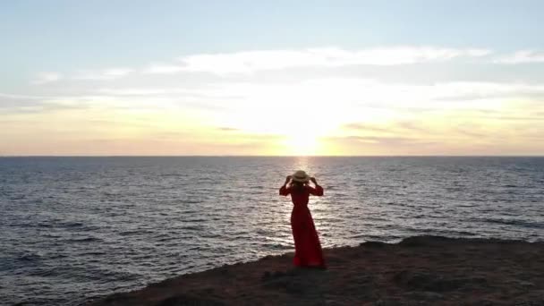 Αεροφωτογραφία μιας νεαρής γυναίκας με κόκκινο φόρεμα και ψάθινο καπέλο περπατά στην άκρη ενός γκρεμού δίπλα στη θάλασσα μόνη της το ηλιοβασίλεμα — Αρχείο Βίντεο