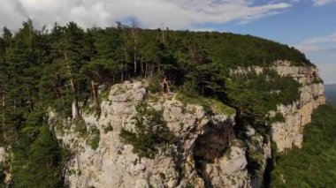 Uzun saçlı, sıska, siyah tişörtlü ve şortlu bir adam uçurumun kenarında duruyor ve etrafındaki orman ve dağlara bakıyor. Hava görünümü