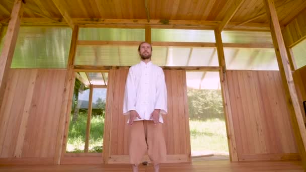 Dlouhovlasý běloch v lehkém a volném oblečení praktikuje qigong tai chi v létě v dřevěné zkušebně. Pomalé pohyby harmonie a klidu — Stock video
