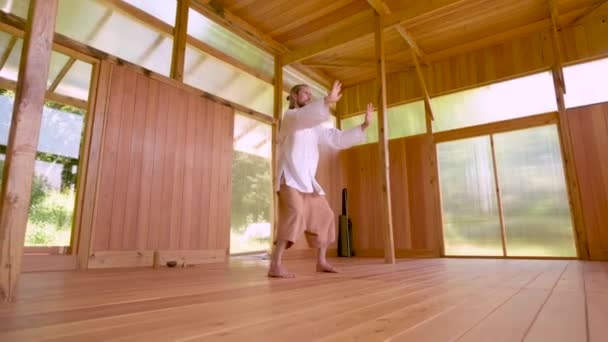 En långhårig vit man i lätt och lös klädsel övar qigong tai chi i ett övningsrum i trä på sommaren. Långsamma rörelser harmoni och lugn — Stockvideo