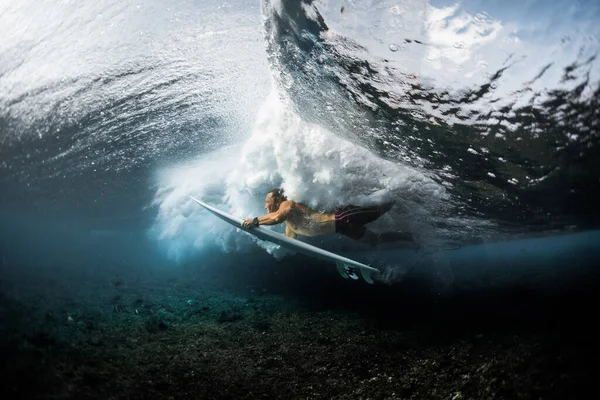 Young surfer dives under the ocean wave with surfboard. Performs trick named Duck Dive to pass the wave