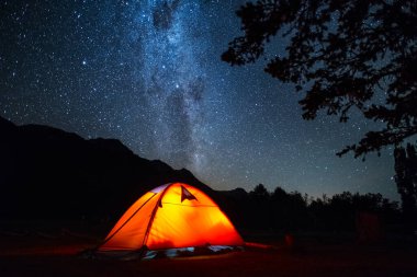 Çadır ve gece gökyüzü. Vurgulanmış turuncu yürüyüş çadırı ve ön planda ağaçlar olan derin yıldızlı gökyüzü..