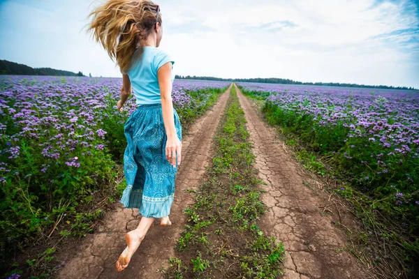 身着连衣裙的女人赤着脚 沿着满是紫色花朵的夏季牧场在乡间小路上奔跑 — 图库照片