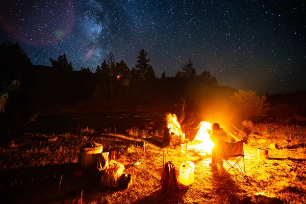 Tourists sit near the fire in the wild area surrounded by pine trees under the starry sky. Image has some noise.