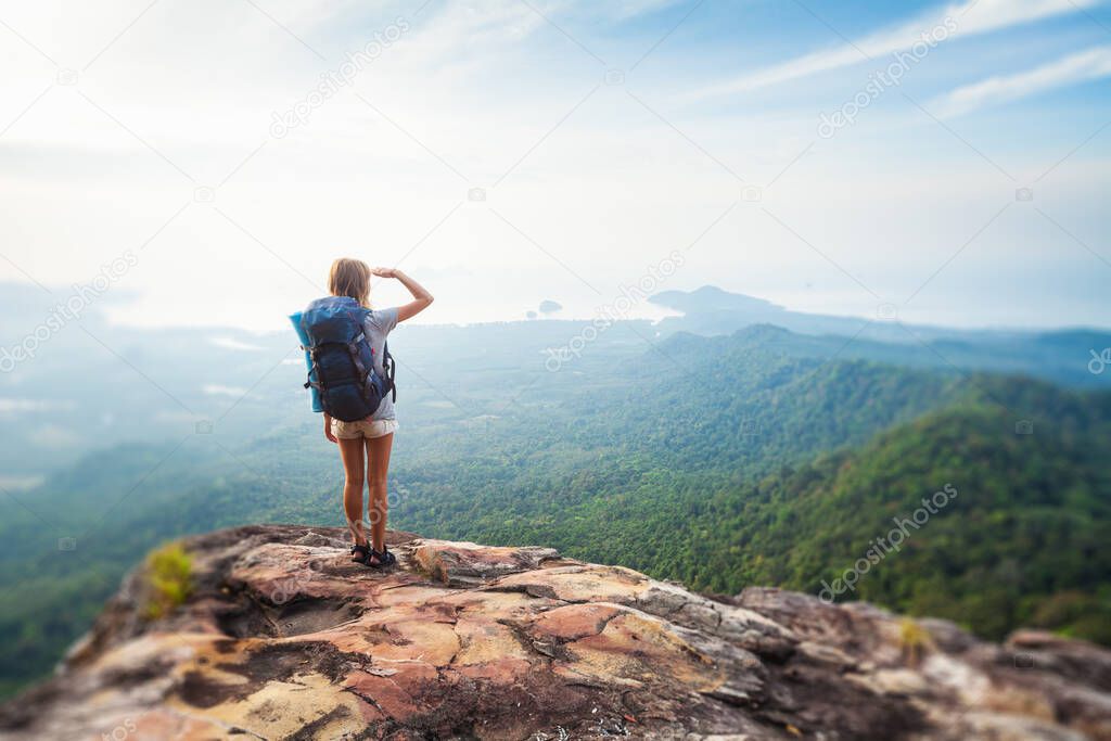 Young woman hiker stands with backpack on the cliff and looks into valley and sea coastline. Tilt shift effect applied