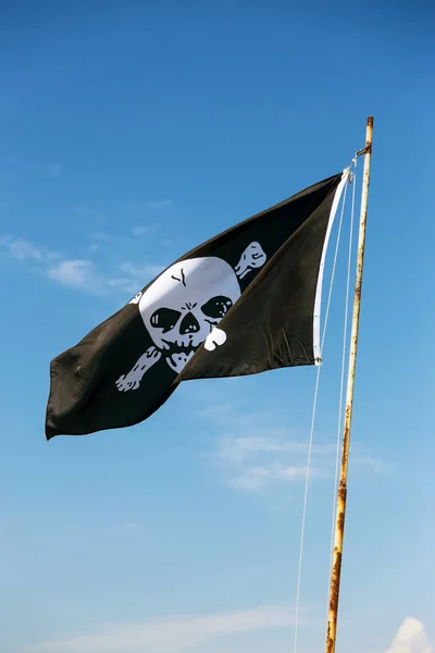 fragment of flag depicting skull as symbol of pirates. Jolly Roger / Skull and Crossbones Pirate flag torn in wind against blue sky. Cheerful Roger or Pirate flag blowing in wind against sky
