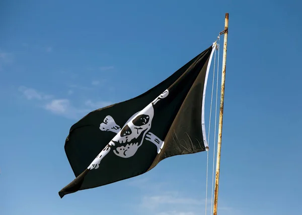 fragment of flag depicting skull as symbol of pirates. Jolly Roger / Skull and Crossbones Pirate flag torn in wind against blue sky. Cheerful Roger or Pirate flag blowing in wind against sky