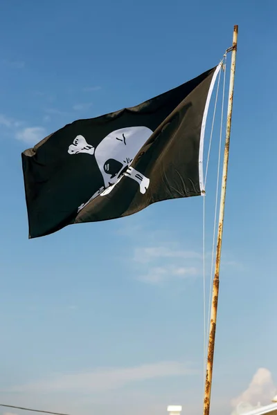fragment of flag depicting skull as symbol of pirates. Jolly Roger / Skull and Crossbones Pirate flag torn in wind against blue sky. Cheerful Roger or Pirate flag blowing in wind against sky