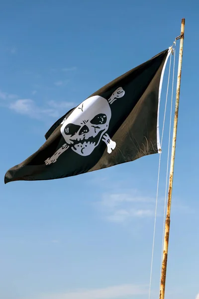 fragment of flag depicting skull as symbol of pirates. Jolly Roger / Skull and Crossbones Pirate flag torn in wind against blue sky. Cheerful Roger or Pirate flag blowing in wind against sky