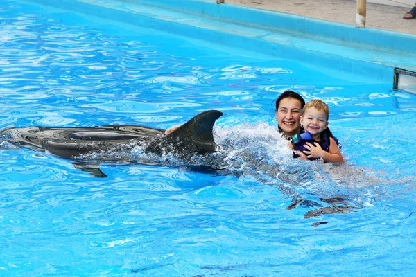 快乐美丽的年轻女子 与一个小的孩子笑着清澈阳光明媚的日子与海豚在蓝色的游泳池里游泳 — 图库照片