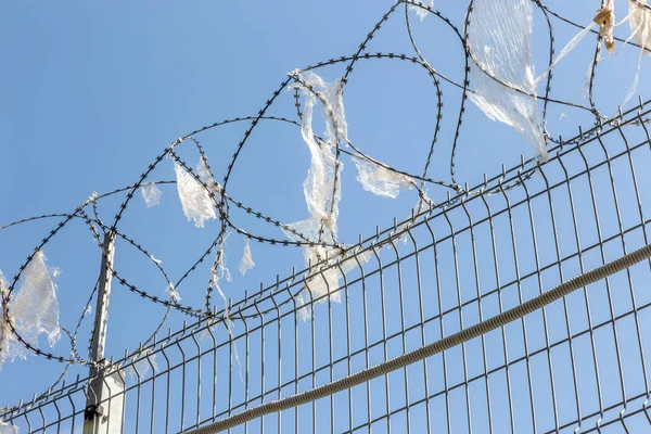 带栅栏的铁丝网 有粘附碎屑 在仓库防护栅栏墙的铁丝网尖顶上 对蓝天的破旧塑料袋 — 图库照片