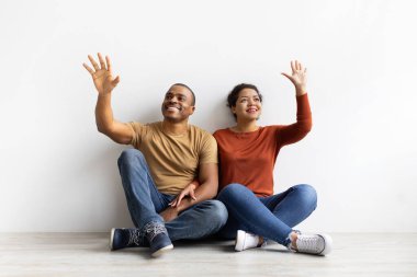 Smiling African American couple sitting on floor, raising hands dreamy, imagining something. Portrait of young man and woman in casual clothes enjoying fun lifestyle moment indoors