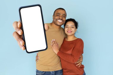 Smiling African American couple hugging while holding smartphone with blank screen. Portrait symbolizing love, partnership, digital lifestyle, and online communication, mockup